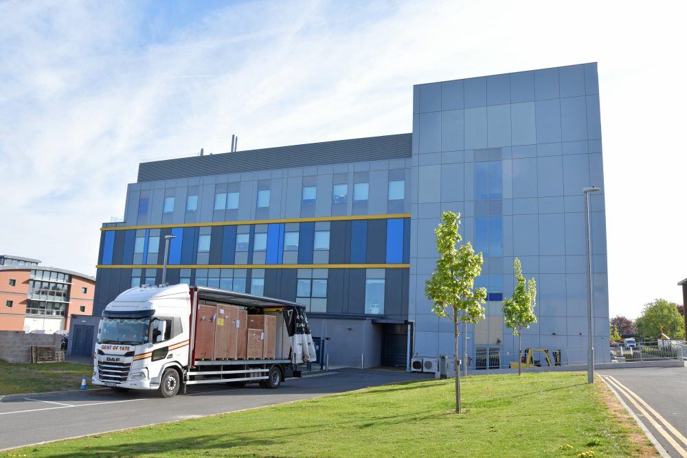 Large lorry with 5 large wooden crates on it outside the Franklin building, a rectangular grey and blue building with a yellow stripe around it.